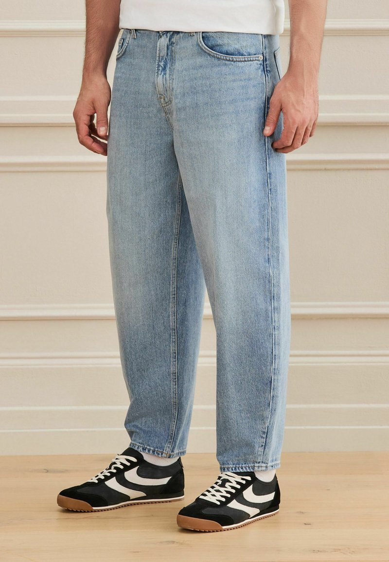 Homme portant un jean bleu clair coupe large et des baskets noires avec des rayures blanches ondulées, debout sur un sol en bois clair.
