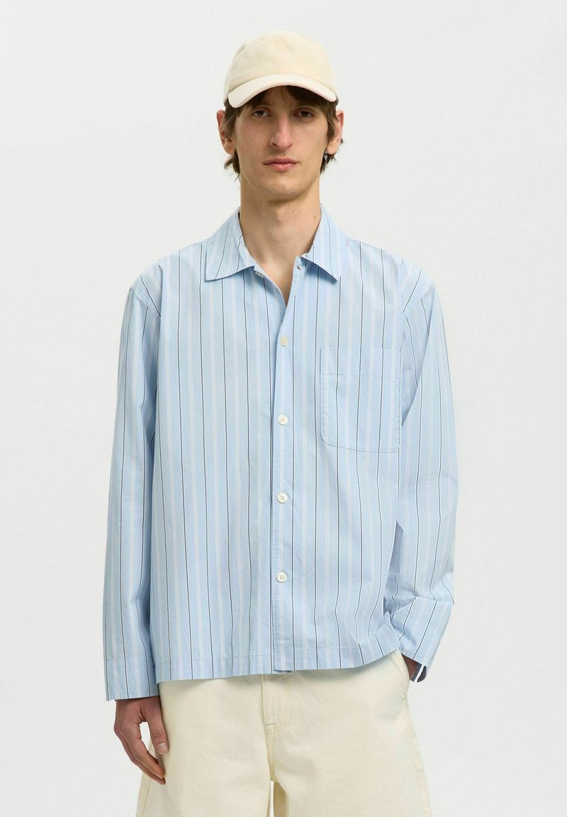 Homme portant une casquette beige, une chemise bleu clair à manches longues avec de fines rayures verticales, et un pantalon écru sur fond uni.
