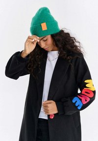 Kobieta w zielonej dzianinowej czapce i czarnym płaszczu z kolorowym napisem „MORE” na rękawie, poprawiająca czapkę jedną ręką.