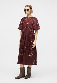 Vrouw met een zonnebril, een bordeauxrode midijurk met geborduurde bloemen en uitgesneden kanten details, gecombineerd met bruine laarzen tot halverwege de kuit.