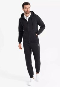 Sort zip-up hættetrøje med snore, forlommer og matchende tapered sweatpants. Båret med hvide sneakers på en ensfarvet baggrund.