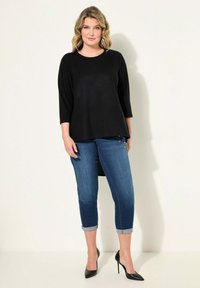 Top negro de manga larga con un dobladillo ligeramente curvado, combinado con jeans azul oscuro remangados en los puños y zapatos de tacón negro.