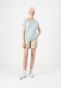 Stribet lyseblå V-hals t-shirt med korte ærmer, parret med beige shorts. Modellen har hvide højhals sneakers og ankelsokker på.