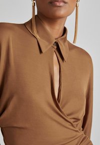 Bruine wikkelshirt met een diepe V-hals, kraag en sleutelgatdetail. Zachte stof met een gladde textuur. Lange, gouden oorbellen zichtbaar.