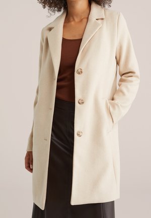 Vrouw draagt een beige jas met knopen over een bruin topje en een zwarte rok, één hand in de jaszak tegen een neutrale achtergrond.