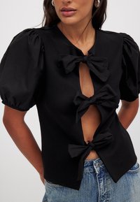 Top negro con mangas abullonadas y tres detalles de lazo en el frente. Presenta un diseño de escote en V profundo y una silueta ajustada.