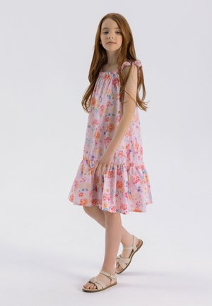 Robe florale rose avec un col smocké et un ourlet à volants. Présente de petits nœuds aux épaules. Portée avec des sandales de couleur claire.