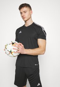 Zwarte sportjersey met een wit drie-strepen ontwerp, gecombineerd met zwarte shorts. Model houdt een kleurrijke voetbals die sterrenpatronen heeft.