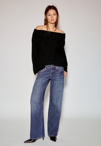 Zwarte gebreide trui met blote schouders, wijde blauwe denim jeans met subtiele glans en zwarte pumps met een spits toelopende neus, getoond tegen een neutrale achtergrond.