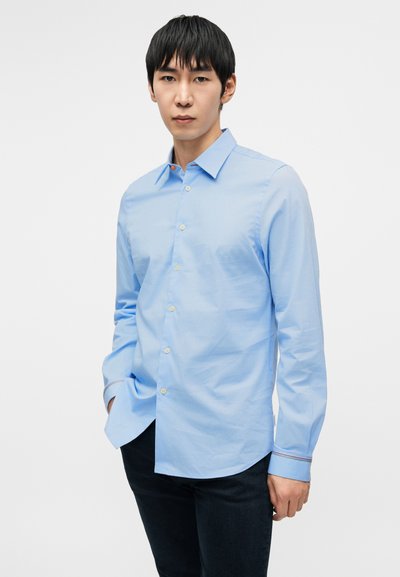 Ανοιχτό μπλε πουκάμισο με μακριά μανίκια, κουμπωτό μπροστινό μέρος, μυτερό γιακά και αντίθετα χρωματιστά διακοσμητικά στα μανίκια. Λεία υφή και εφαρμοστό σχέδιο.