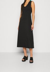 Musta hihaton midi-mekko, jossa on tiukka bodice ja virtaava hame, valmistettu sileästä kankaasta. Yhdistetty paksuihin mustiin platform-sandaaleihin.