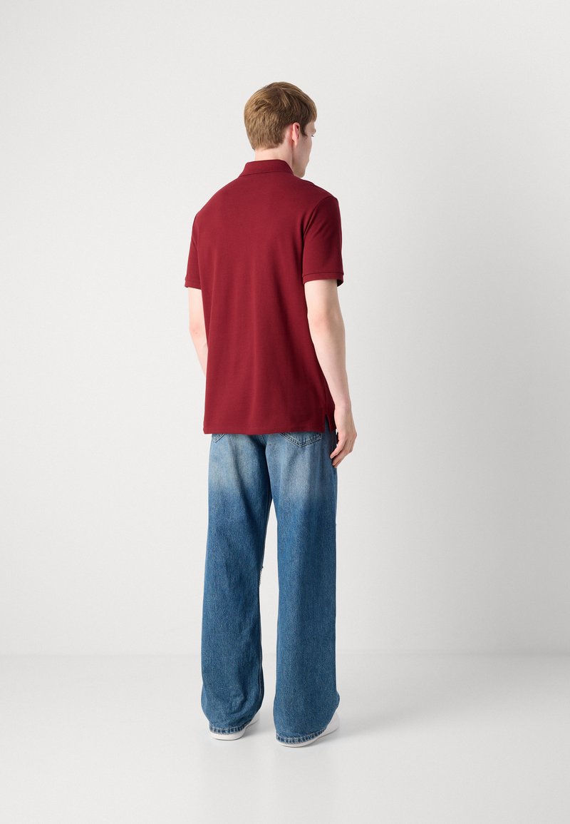 Mężczyzna odwrócony plecami, ubrany w polo z krótkim rękawem w odcieniu bordowym oraz luźne, dwukolorowe niebieskie dżinsy, na jednolitym białym tle.