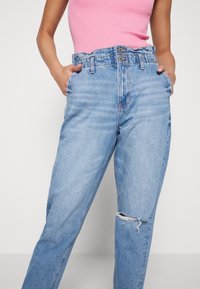 Vaaleansiniset korkeavyötäröiset farkut, joissa on rento leikkaus, ja joissa on kuluneet polvet sekä vyölenkki. Edessä on kaksi taskua ja vetoketju.