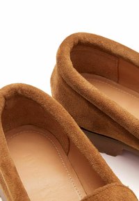 Brązowe zamszowe mokasyny z zaokrąglonymi brzegami, gładkimi skórzanymi wkładkami i widocznymi detalami szycia. Miękka tekstura i casualowy design.