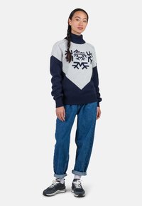 Navy und grauer Strickpullover mit hohem Kragen, ausgestattet mit Schneeflockenmuster und Chevron-Design; kombiniert mit lockeren blauen Jeans und Sneakern.