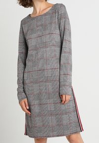 Robe à manches longues avec un motif à carreaux aux teintes noir, blanc et rouge. Comprend un col bateau et des fentes latérales avec des accents rouges.