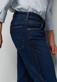 Donkerblauwe denim jeans met een aansluitend design, voorzien van gele stiksels, twee achterzakken en een zichtbaar leren label op de tailleband.