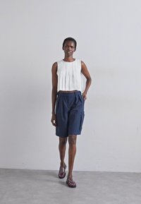 Weißes, plissiertes Cropped-Top kombiniert mit dunkelblauen, gestreiften Shorts. Das Modell trägt dunkle Slipper. Einfache, klare Linien und weiche Texturen.