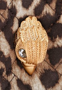 Gouden metalen sluiting in de vorm van gelaagde bladeren met een klein kristaldetail, tegen een gequilt, luipaardpatroon achtergrond.