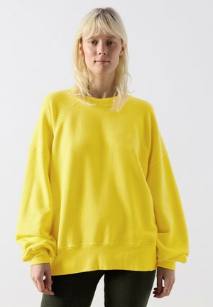 Blonde woman wearing loose, bright yellow long-sleeve sweatshirt and dark pants, standing against a plain white background.