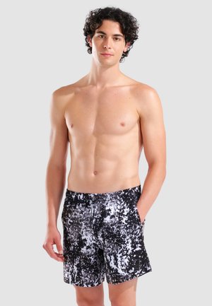Junger Mann steht oberkörperfrei, trägt schwarz-weiße Badeshorts mit abstraktem Muster und hat die Hände in den Taschen.