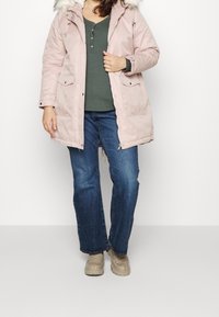 Eine Person steht da und trägt eine pinke Parka mit einer fellgefütterten Kapuze, ein grünes Oberteil, blaue Jeans und beige Schuhe vor einem schlichten weißen Hintergrund.
