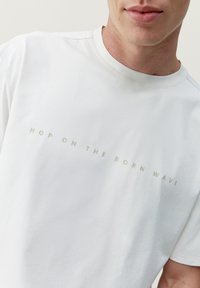 Biela bavlnená tričko s krátkymi rukávmi, s okrúhlym výstrihom a predným potlačeným textom "NASKOČ NA VLNIU ZRODENIA" v svetlošedej farbe.