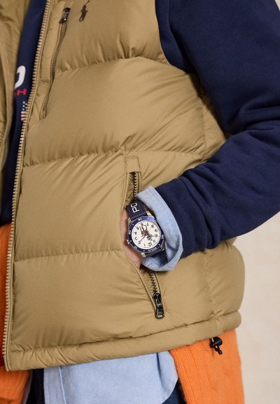 Ręka w kieszeni beżowej kamizelki puchowej, nosząca granatowy zegarek z białą tarczą i logo Polo, ułożona na niebieskiej koszuli i granatowym swetrze.