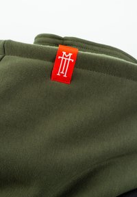 Olive green fabric with a smooth texture; features a red label with white logo. Clean edges and minimal design.