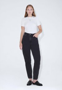 Hochtaillierte schwarze Jeans mit schmalem Schnitt, strukturierten Denim, kombiniert mit einem eng anliegenden weißen T-Shirt und schwarzen Slipschuhen.