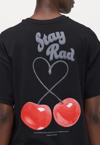 Melna kokvilnas t-kreklu ar lielu grafiku, kurā ir savītas sarkanas ķirši un teksts "Stay Rad" virsū. Gluda tekstūra ar košām krāsām.