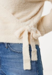 Cremfarbener, fuzzy Pullover mit Wickeldesign und einer Bindeschließe an der Taille, kombiniert mit blauen Jeans. Die weiche Textur ist sichtbar.