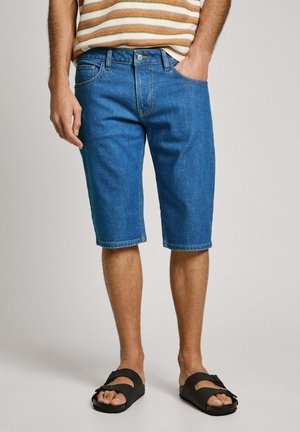Jeansshorts in een middelblauwe tint, met een klassieke pasvorm, vijf zakken en stikdetails. Draagt met zwarte slidesandalen.
