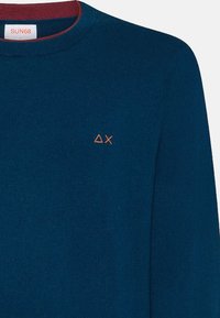 Blauer Pullover mit rundem Ausschnitt und burgunderfarbenen Akzenten. Verfügt über ein kleines oranges Logo auf der linken Brust. Hergestellt aus weichem, strukturiertem Stoff.
