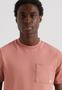 Korte mouwen, ronde hals t-shirt in lichtroze stof met een borstzak, een soepele textuur en een klein label op de zak.