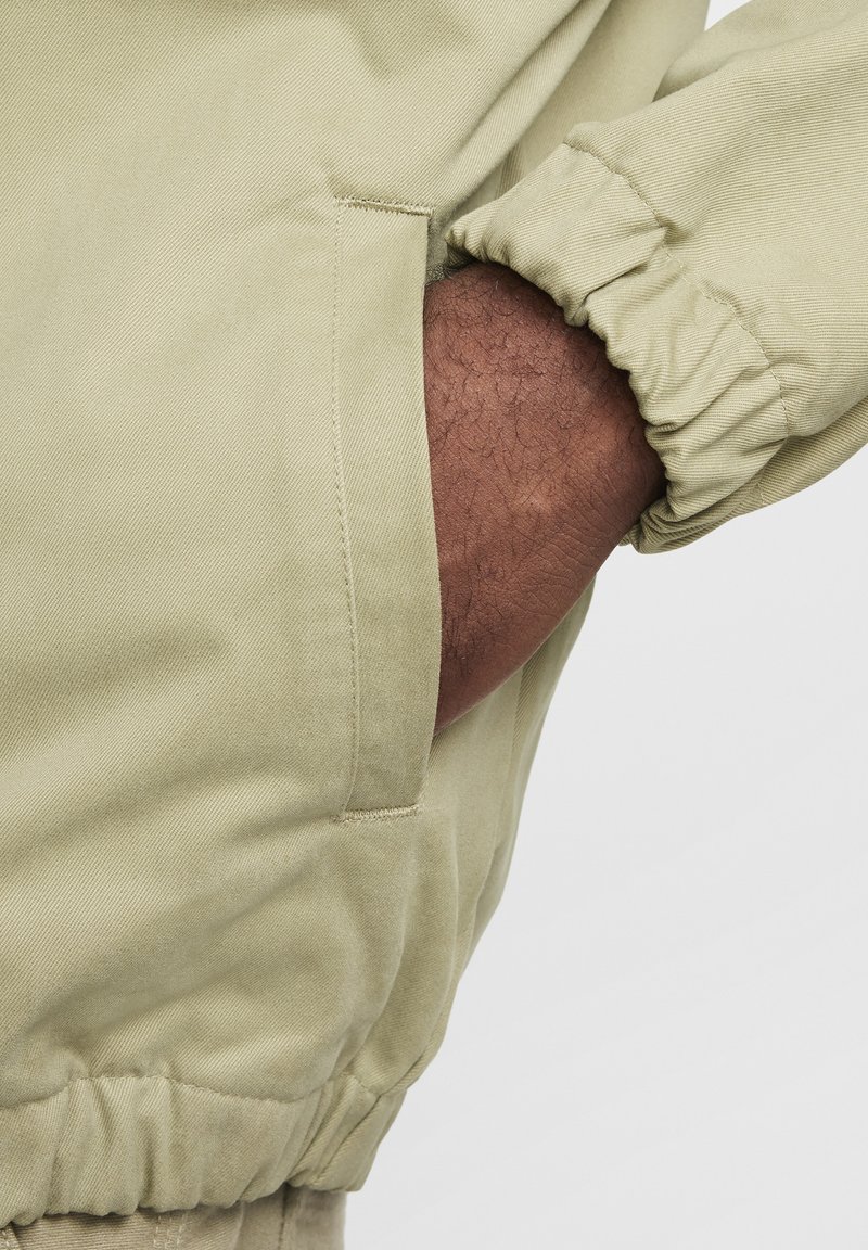 Veste vert olive clair avec des poignets élastiques et une poche frontale. Le tissu est texturé et semble durable, adapté à une tenue décontractée.