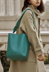 Borsa a secchiello in pelle verde acqua con logo dorato, dotata di una tracolla staccabile e una superficie liscia. Indossata con un cappotto beige.