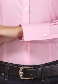Camicia button-up rosa chiaro dalla texture liscia, con bottoni bianchi, indossata con una cintura in pelle nera sopra jeans in denim blu.