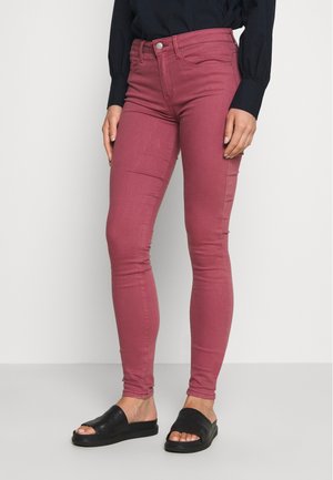 Jeans ajustados de color burdeos, hechos de material elástico. Con cintura alta y diseño de cinco bolsillos. Combinados con sandalias tipo slide negras.
