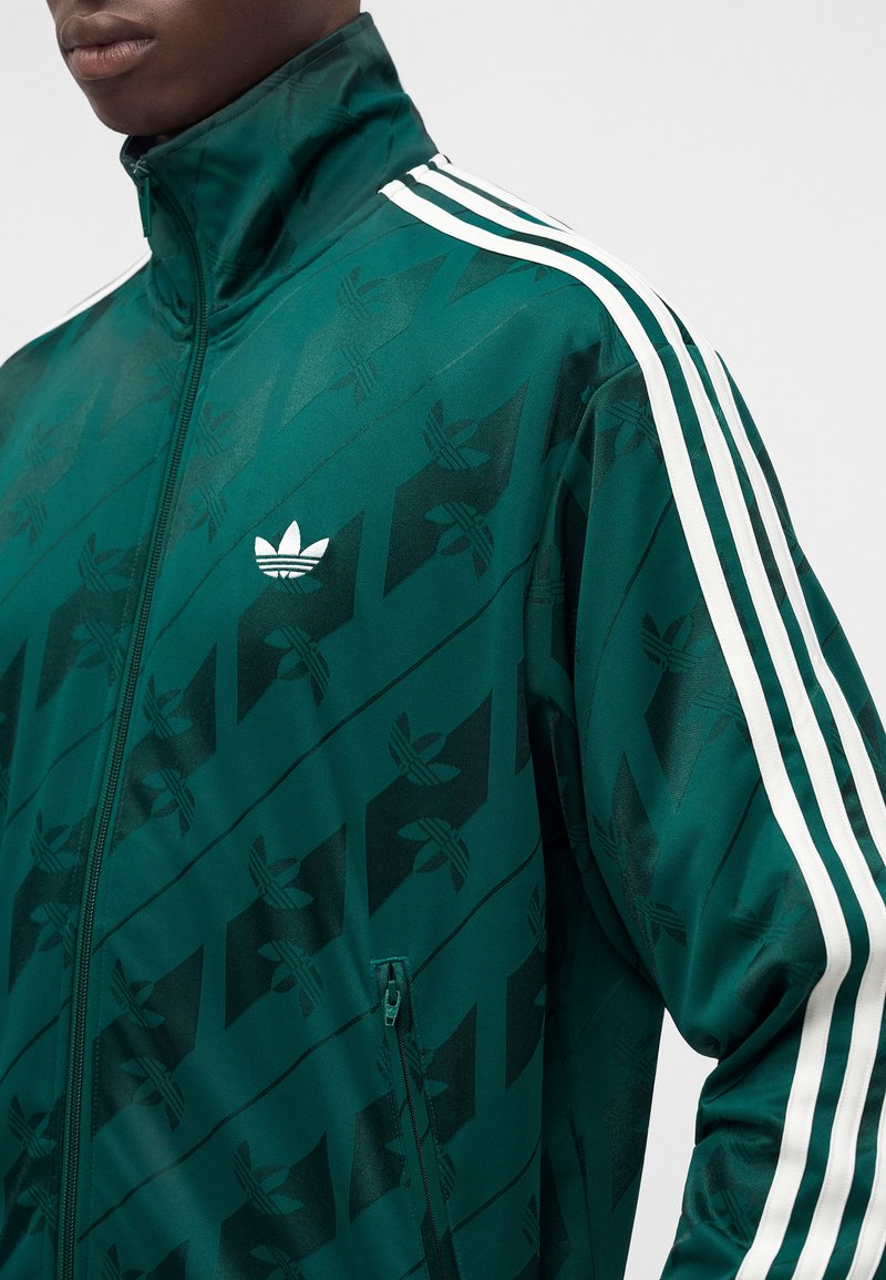 Persona vistiendo una chaqueta de chándal Adidas de color verde oscuro con rayas blancas en las mangas y un bolsillo con cremallera, sobre un fondo liso.