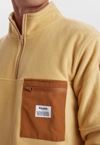 Fleece pullover in zacht geel met een kwart-rits hals, voorzien van een bruine rits borstzak en een label op de zak.