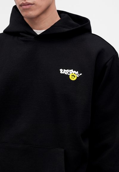 Homme portant un sweat à capuche noir avec un texte blanc et un petit smiley jaune sur la poitrine.