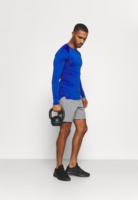 Langärmliges, enganliegendes blaues Sporthemd mit grauen Shorts, kombiniert mit schwarzen Sportschuhen. Hält eine schwarze Kettlebell mit blauen Akzenten.