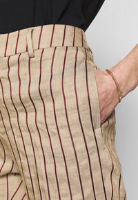 Beżowe spodnie w paski z wąskimi, czerwonymi liniami. Posiadają dopasowany pas, kieszenie boczne i teksturowaną tkaninę.