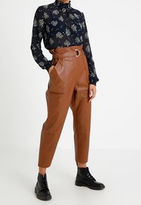 Pantalon taille haute en similicuir marron avec boucle de ceinture, associé à un chemisier à manches longues bleu marine à imprimé floral et des bottines noires à lacets.