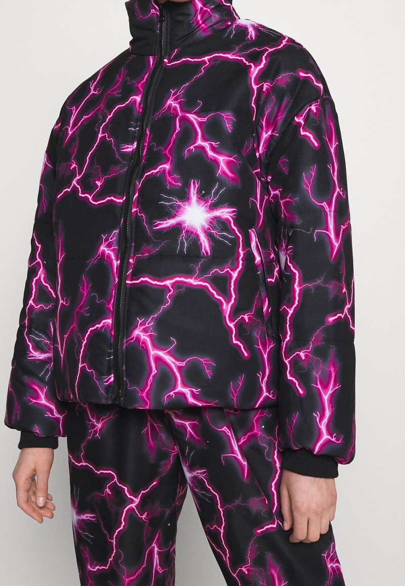 Doudoune noire et pantalon avec des motifs d’éclairs rose vif et une fermeture éclair frontale, porté par une personne avec des mains visibles.