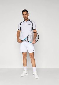 Λευκό polo με κοντά μανίκια και μαύρες λεπτομέρειες, με γιακά και φερμουάρ, συνδυασμένο με λευκά σορτς και αθλητικά παπούτσια, κρατώντας ρακέτα τένις.