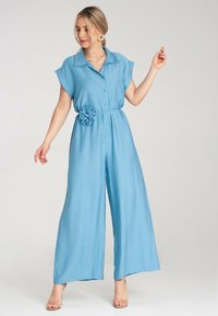 Jasnoniebieski, krótki kombinezon z kołnierzykiem, z przodu zapinany na guziki, z tkaninowym kwiatem jako akcentem w pasie. Luźny, szeroki krój nogawek.