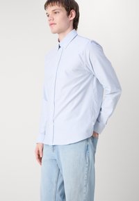 Chemise boutonnée rayée bleue et blanche, à manches longues, avec un détail de col. Portée avec un jean bleu clair, mettant en avant une tenue décontractée.