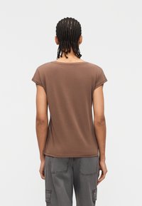 Άτομο με πλεξούδες που κοιτάζει προς τα πίσω, φοράει καφέ κοντομάνικο πουκάμισο και γκρι παντελόνι cargo μπροστά σε απλό ανοιχτό φόντο.